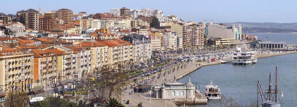 Vista de edificios con fachadas de distintos colores junto a un paseo, un puerto con embarcaciones y una amplia superficie de agua.